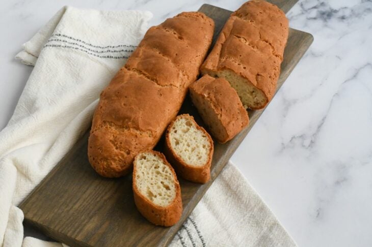 two vegan baguettes on a wooden cutting board, with one loaf partially sliced