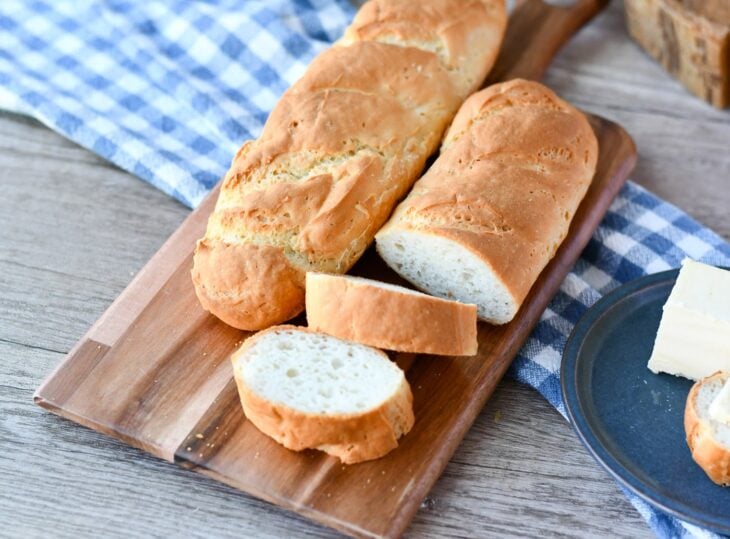 two vegan baguettes on a wooden cutting board, with one partially sliced